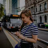 Woman waiting at a tram stop in Melbourne, smiling at her phone, wearing white headphones